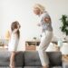 Cute little girl having fun playing with smiling grandmother jumping on couch together, happy granny and active kid grandchild dancing on sofa, grandma and granddaughter laughing playing at home