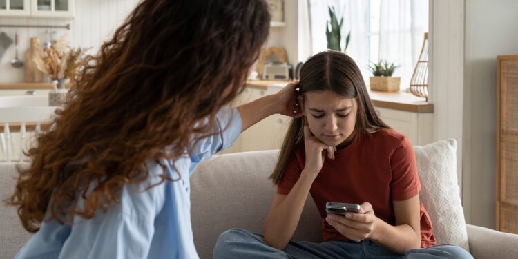 Supportive mother comforting sad teenage girl rejected by friends, helping depressed teen daughter to deal with breakup. Upset depressed adolescent child looking at phone at home, waiting for call