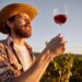 Low angle of cheerful male oenologist standing with glass of red wine in garden in sunset and digestating tasty drink