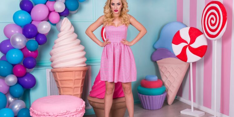 Portrait of joyful young woman in pink dress on background decorated with huge candies and ice cream. Barbie
