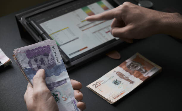 Male had holding colombian banknotes while he does the math on a tablet
