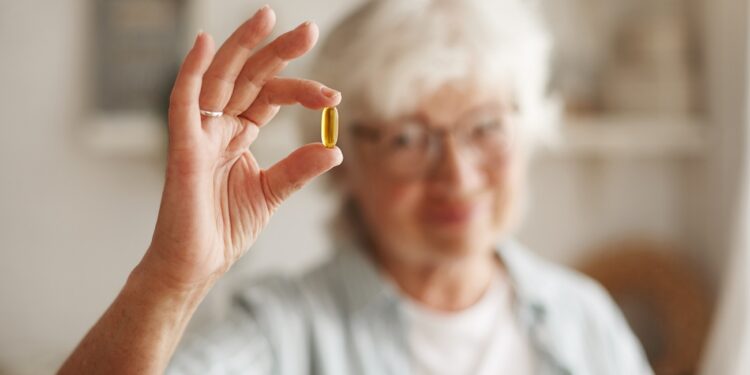 Food, nutrition, diet and health concept. Close up shot of elderly woman's hand holding fish oil or omega-3 polyunsaturated fatty acid supplement in shape of capsule, going to take one during lunch