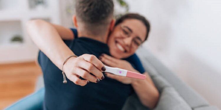 A young Caucasian woman is cheerfully hugging her husband and holding a positive pregnancy test in the forefront.