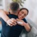 A young Caucasian woman is cheerfully hugging her husband and holding a positive pregnancy test in the forefront.