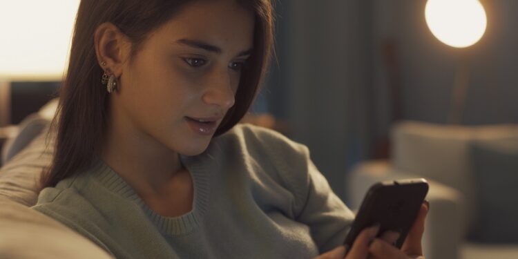 Smiling teenager relaxing on the sofa and connecting online with her smartphone, she is social networking and chatting