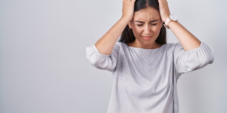 Young hispanic woman standing over white background suffering from headache desperate and stressed because pain and migraine. hands on head.
