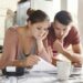 Worried young Caucasian married couple reading important notification from bank while managing domestic finances and calculating their expenses at kitchen table, using laptop computer and calculator