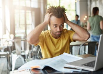 International Afro American student feeling stressed, keeping hands on his head, staring at laptop screen in frustration and despair after he made serious mistake in calculation, working on research