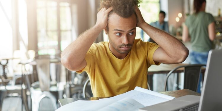 International Afro American student feeling stressed, keeping hands on his head, staring at laptop screen in frustration and despair after he made serious mistake in calculation, working on research