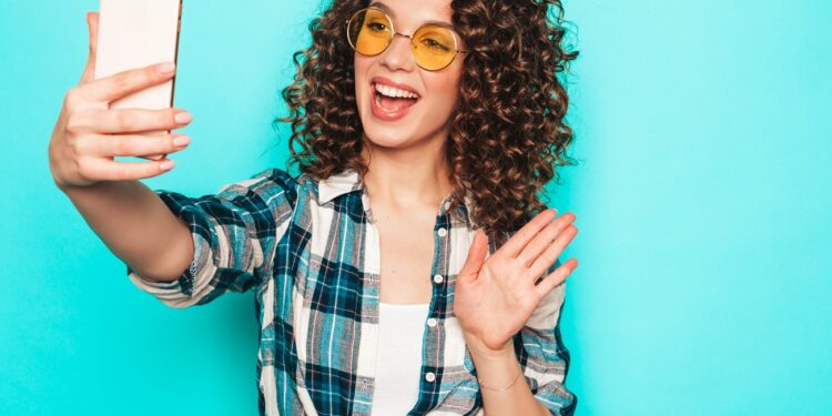 Portrait of beautiful smiling model with afro curls hairstyle dressed in summer hipster clothes.Sexy carefree girl posing in studio near blue wall.Trendy funny and positive woman makes selfie