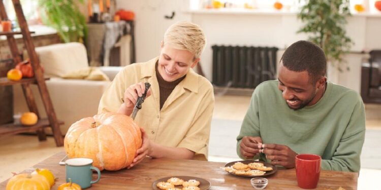 Positive young multi-ethnic friends sitting at wooden table and enjoying Halloween preparation: Black man decorating cookies while blond girl carving pumpkin; Shutterstock ID 2161486659; purchase_order: 09262022; job: Editorial Associate; client: Ellie Spencer-Failes; other: