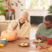 Positive young multi-ethnic friends sitting at wooden table and enjoying Halloween preparation: Black man decorating cookies while blond girl carving pumpkin; Shutterstock ID 2161486659; purchase_order: 09262022; job: Editorial Associate; client: Ellie Spencer-Failes; other: