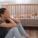 Side view of mother covering face crying from exhaustion sitting on floor next to baby crib while child napping, can't handle difficulties of maternity, having postnatal depression symptoms