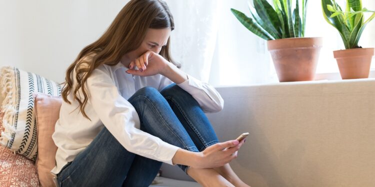 Young woman cry reading sms on smartphone suffer from breakup with boyfriend. Frustrated female in tears on couch with mobile phone in hand get message with bad news, betrayal from friend or accident