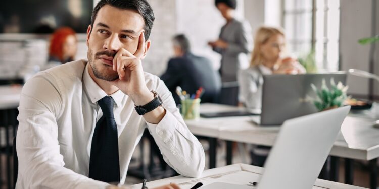 Pensive businessman sitting at desk in the office and thinking of new ideas. There are people in the background.