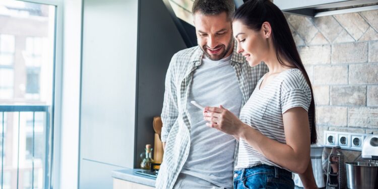 smiling couple using smartphone together in kitchen, smart home concept