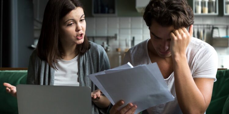 Angry wife scolding blaming upset husband of unpaid bills, bankruptcy or past due debt, unhappy millennial couple having financial problems arguing at home sitting on sofa with laptop and papers