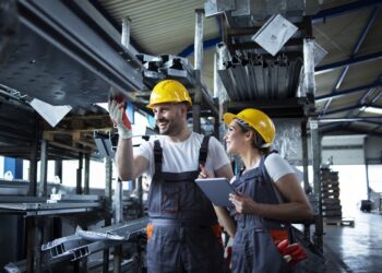 Factory workers checking inventory with tablet computer in industrial warehouse full of metal parts.