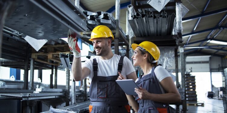 Factory workers checking inventory with tablet computer in industrial warehouse full of metal parts.