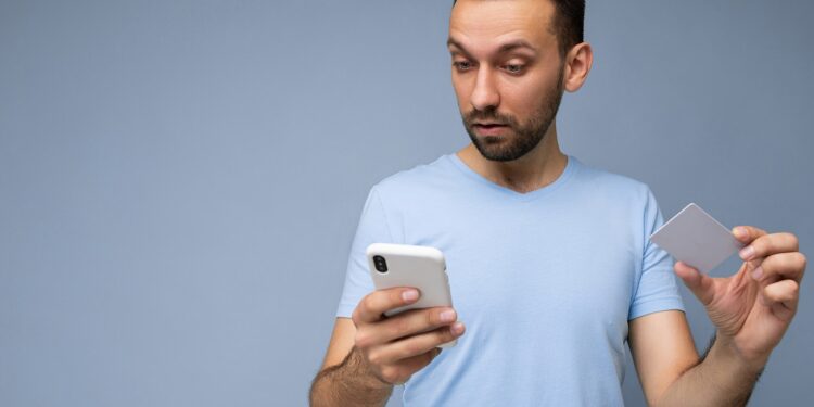 Handsome brunet man wearing everyday clothes isolated on blue background wall with copy space holding and using mobile phone and plastic credit card making payment.