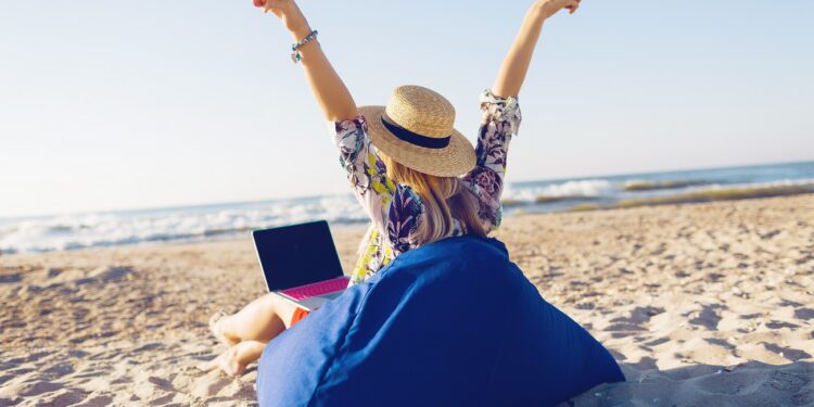 Beautiful young woman working with laptop on the tropical beach. Happy traveling woman in straw hat using laptop on holidays.