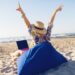 Beautiful young woman working with laptop on the tropical beach. Happy traveling woman in straw hat using laptop on holidays.