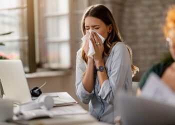 Young sick businesswoman sneezing in a tissue while working in the office.