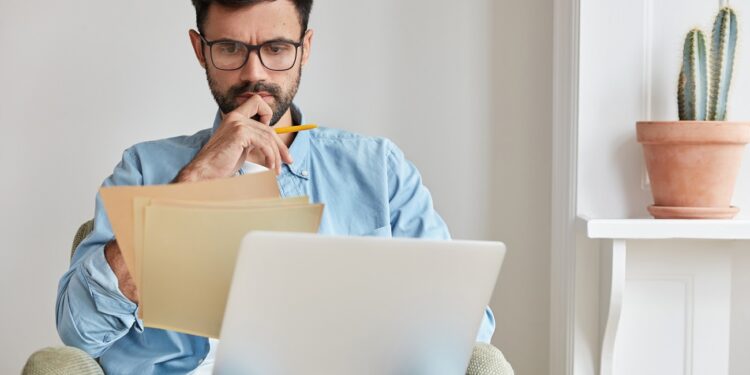 Serious concentrated businessman in spectacles for vision protection, works on laptop computer, studies documents, does distance work at home interior. Male financier works on business report