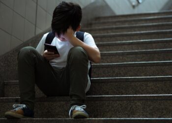 Young teenage Asian student boy sit on stairs alone holding smartphone and cover his face in despair and frustration at school for social media cyberbullying concept