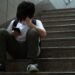 Young teenage Asian student boy sit on stairs alone holding smartphone and cover his face in despair and frustration at school for social media cyberbullying concept