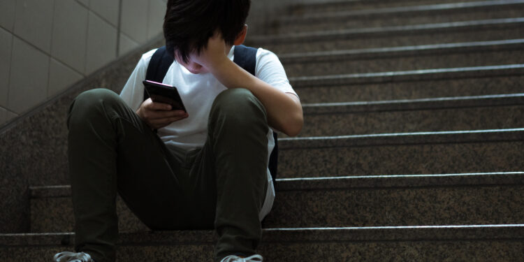 Young teenage Asian student boy sit on stairs alone holding smartphone and cover his face in despair and frustration at school for social media cyberbullying concept