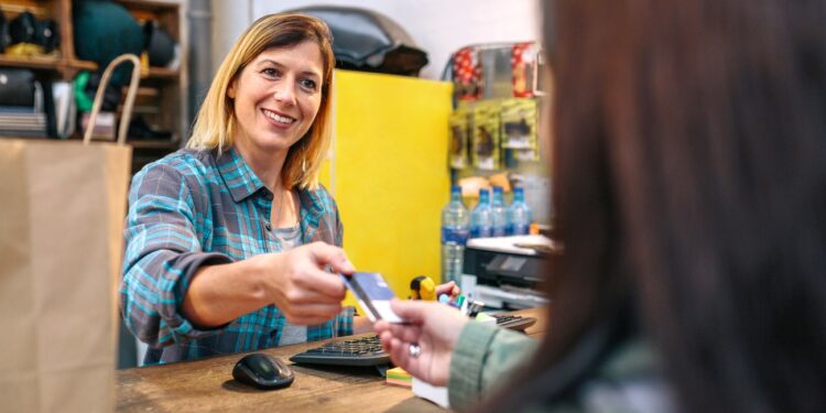 Smiling blonde woman shop assistant taking credit card of client to charge purchase. Cheerful female cashier serving to woman customer in vintage store. Small business and local commerce concept.