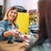 Smiling blonde woman shop assistant taking credit card of client to charge purchase. Cheerful female cashier serving to woman customer in vintage store. Small business and local commerce concept.