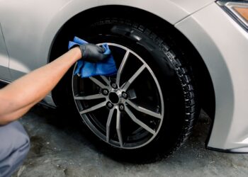 Car rims cleaning, car detailing wash concept. Cropped close up photo of male hand in black rubber glove with blue microfiber cloth washing car alloy wheel at car wash service.