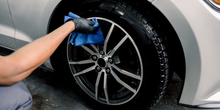 Car rims cleaning, car detailing wash concept. Cropped close up photo of male hand in black rubber glove with blue microfiber cloth washing car alloy wheel at car wash service.