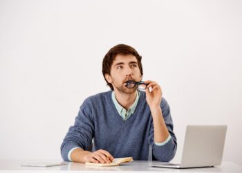 Handsome wondering concentrated young businessman sitting at the office desk writing down something to notebook with a pencil. Looking away. Isolated on white background.