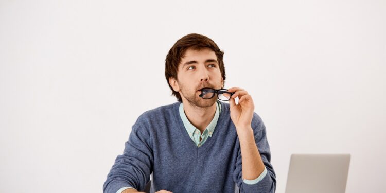 Handsome wondering concentrated young businessman sitting at the office desk writing down something to notebook with a pencil. Looking away. Isolated on white background.