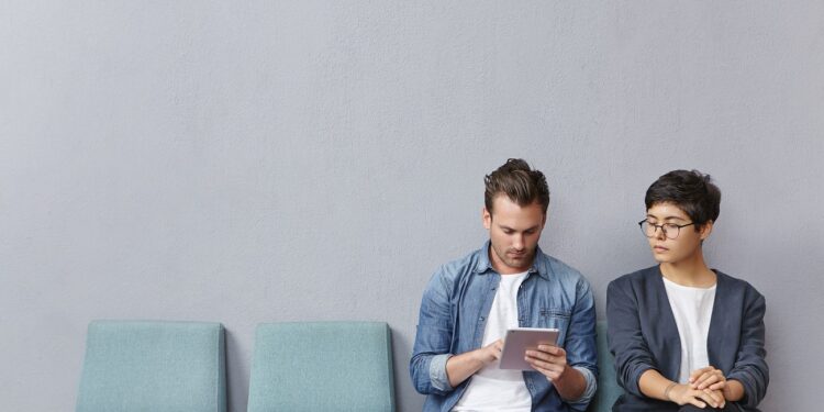 Stylish man using tablet computer whle sitting in queue for doctor and young woman looking at what he is doing. Two people sitting on chairs, using free internet connection while working with gadget