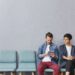 Stylish man using tablet computer whle sitting in queue for doctor and young woman looking at what he is doing. Two people sitting on chairs, using free internet connection while working with gadget