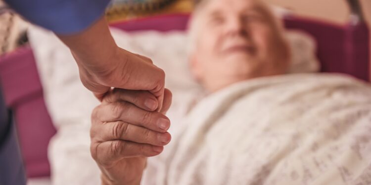 Cropped image of old man looking at nurse, holding her hand and smiling while lying in bed in the hospital ward