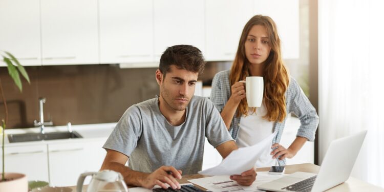 Candid shot of young American couple managing family budget, reviewing bank accounts using laptop and calculator in kitchen. Man and woman doing paperwork together, paying taxes online on notebook pc