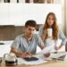 Candid shot of young American couple managing family budget, reviewing bank accounts using laptop and calculator in kitchen. Man and woman doing paperwork together, paying taxes online on notebook pc