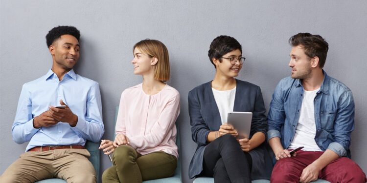 Horizontal portrait of people sit in queue, have pleasant conversation with each other, share ideas and life experience, isolated over grey concrete wall. Diverse group in row, speak and hold gadgets