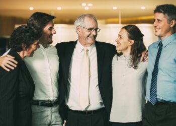 Closeup of smiling senior business man embracing colleagues. They are standing and with blurred view in background.