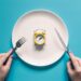 Hands holding knife and fork above alarm clock on a plate on blue background. Intermittent fasting, Ketogenic dieting, weight loss, meal plan, and healthy food concept.