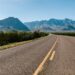 A beautiful shot of an empty road in the middle of plants and mountains in the distance with a clear sky in the background