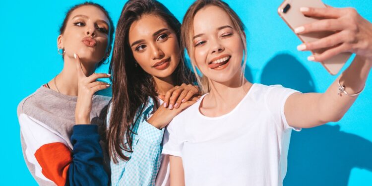 Three young smiling hipster women in summer clothes.Girls taking selfie self portrait photos on smartphone.Models posing near blue wall in studio.Female showing positive emotions