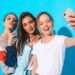 Three young smiling hipster women in summer clothes.Girls taking selfie self portrait photos on smartphone.Models posing near blue wall in studio.Female showing positive emotions