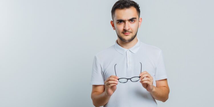 young male holding glasses in t-shirt, pants and looking confident. front view.