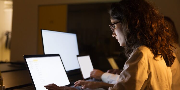 Business people using computers in dark office. Professional male and female business colleagues using laptops with illuminated screens late at night. Working late concept